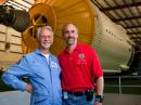 Owen Garriott, W5LFL (left) and his son Richard, W5KWQ, stand next to the simulator at the Johnson Space Center in Texas. Owen made history in 1983 as the first ham to make QSOs from space. Richard hopes to follow that first as he makes his own QSOs on his own voyage to space, set to begin on October 12. [Andrew Yates, Photo]